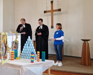 Zwei Menschen in geistlicher Kleidung und ein Mensch in Jeans und blauem Shirt vor einem Kreuz