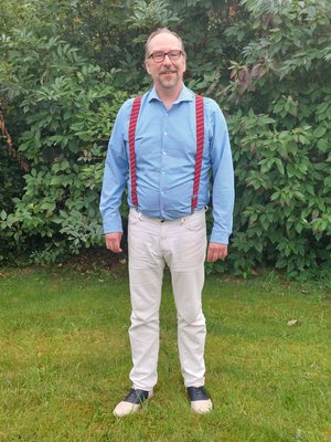Ein Mann in heller Hose und blauem Hemd sowie roten Hosenträgern sowie einer Brille lächelt in die Kamera