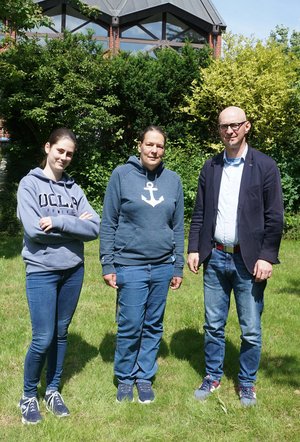 Zwei Frauen und ein Mann stehen in einem Garten, im Hintergrund eine Kirche