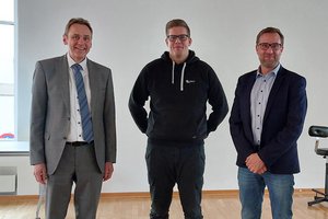 Drei Männer, davon zwei in Anzügen, lächeln in die Kamera